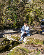 Une femme assise sur un rocher au bord d'une rivière rit tandis qu'un garçon court hors champ.