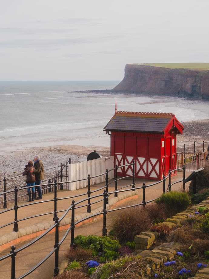 Coastal scene with red beach hut, people on walkway, cliffs, and gardens overlooking a sandy bay.