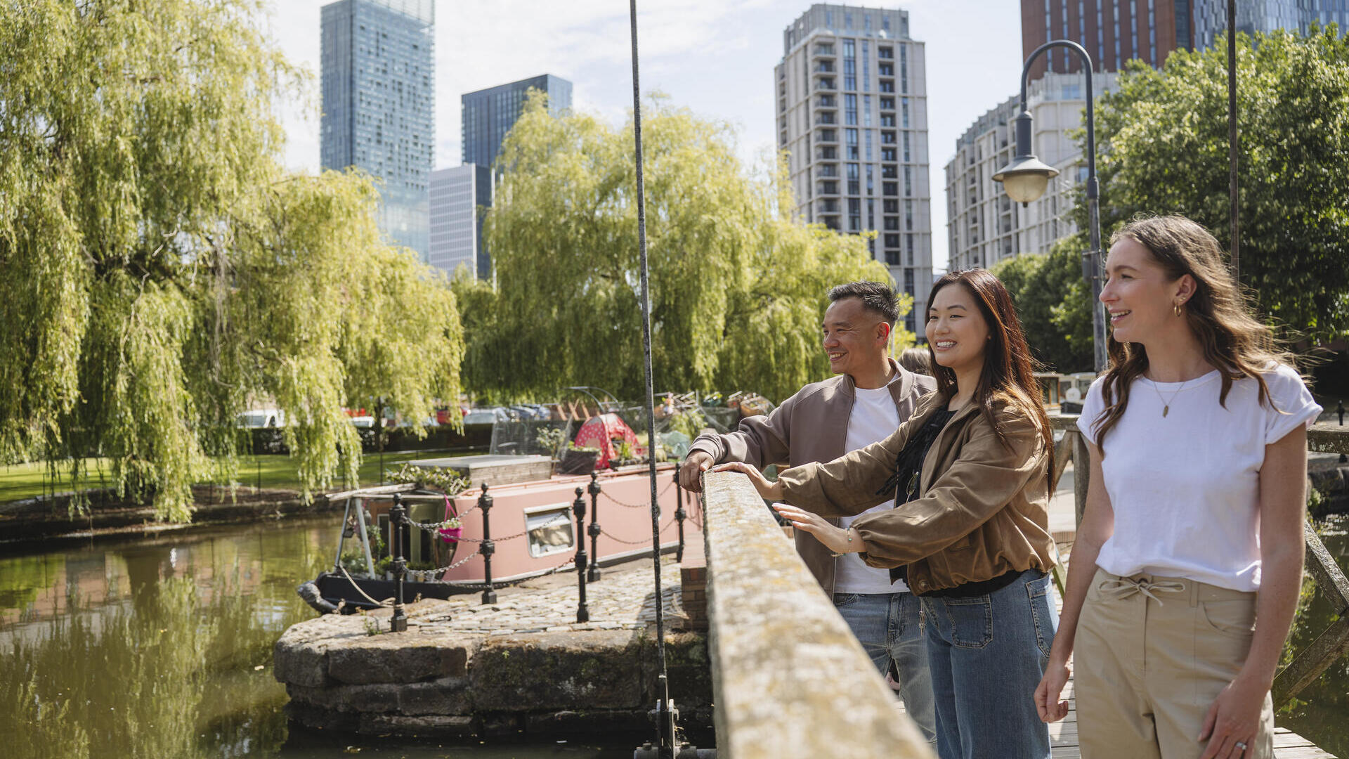 Three people stand on a canal bridge with modern high-rise buildings, green trees, and a pink canal boat in the background.