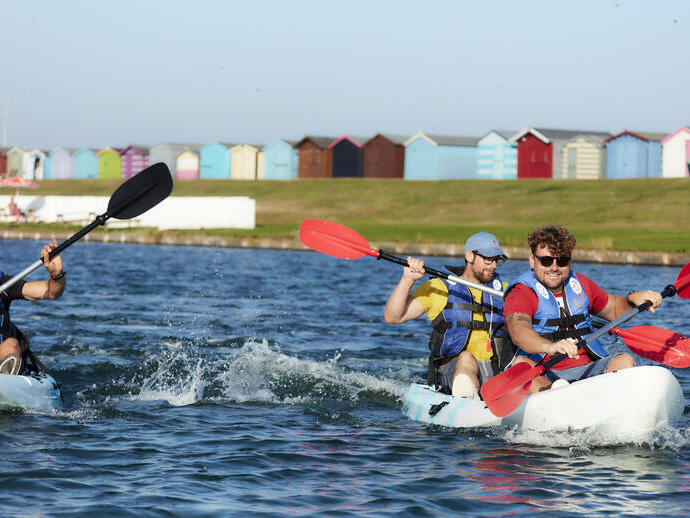 Amigos practicando kayak en la isla de Mersea con cabañas de playa al fondo