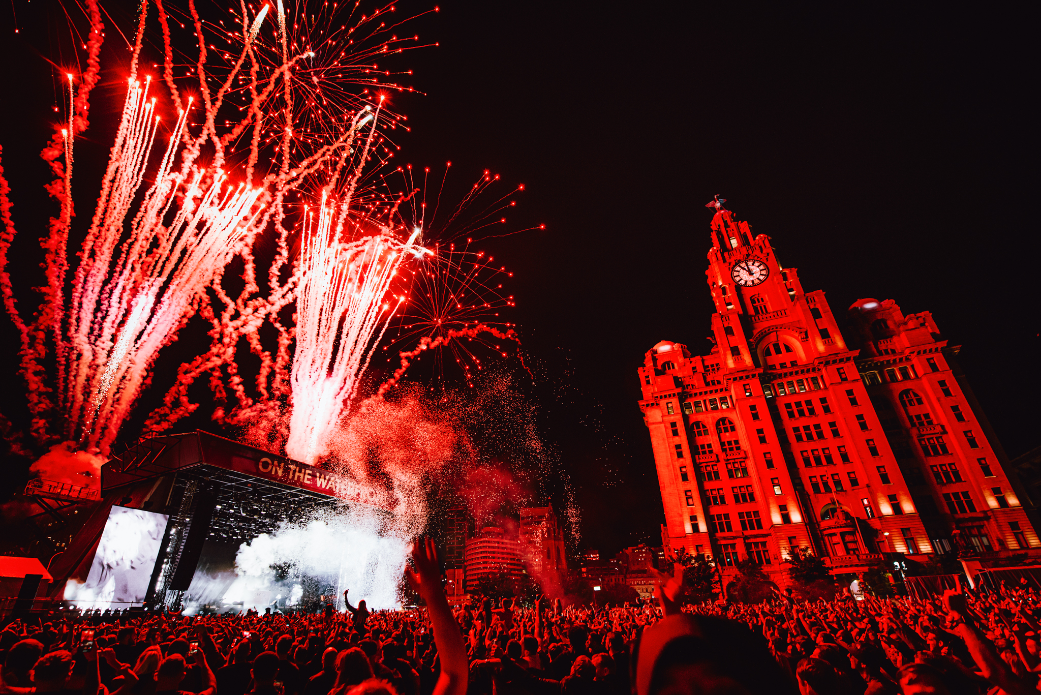 Crowds of people watching a band at a music festival as fireworks explode, in front of the Liver Building.