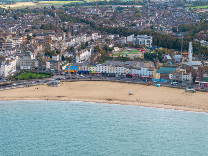 Aerial view of a sandy beach with a promenade, amusement rides, and a town with buildings and greenery in the background.