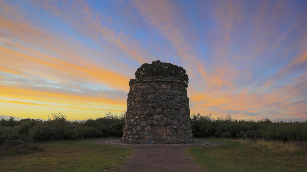 A small stone tower with a plaque on it surrounded by grass and moor under a vibrant blue and orange sky at sunset.