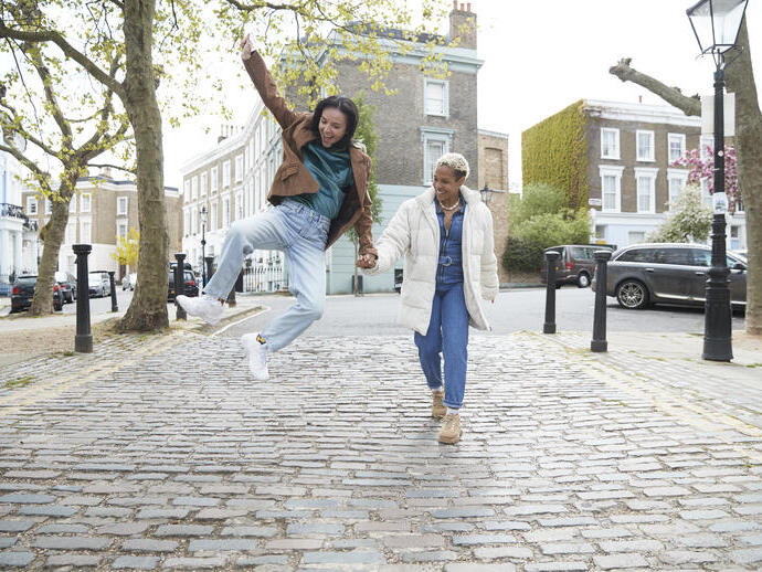 Happy young woman jumping while holding girlfriend's hand walking on a footpath in a street