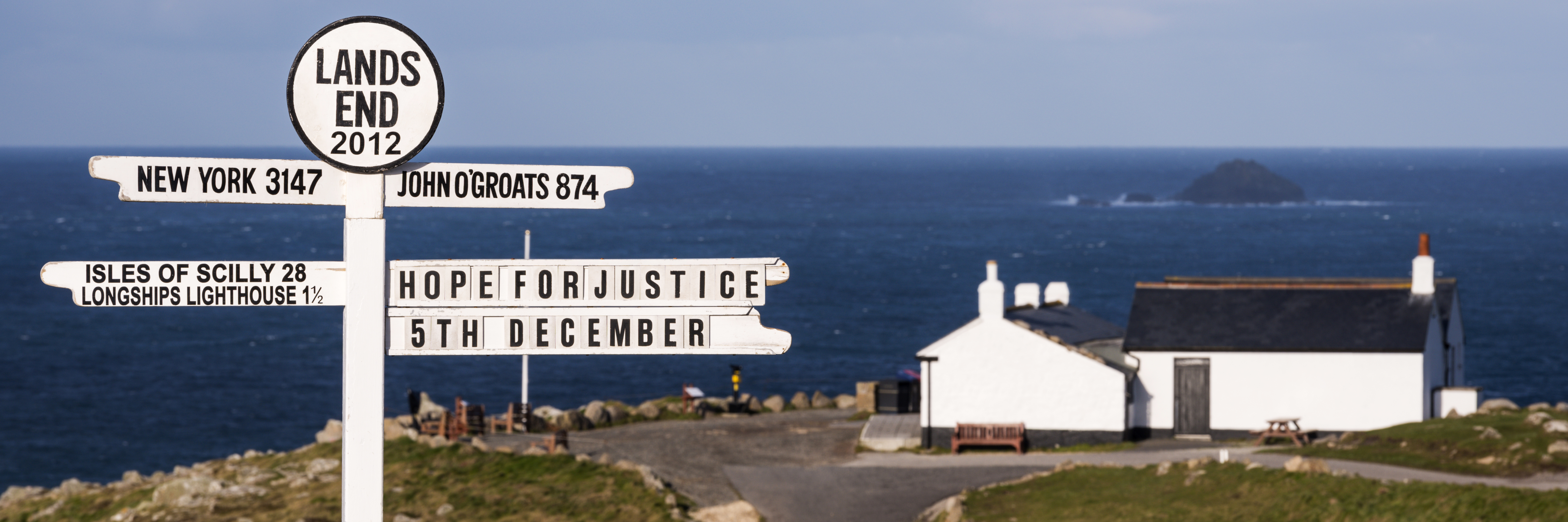 White signpost on a clifftop with the sea and a white building in the distance