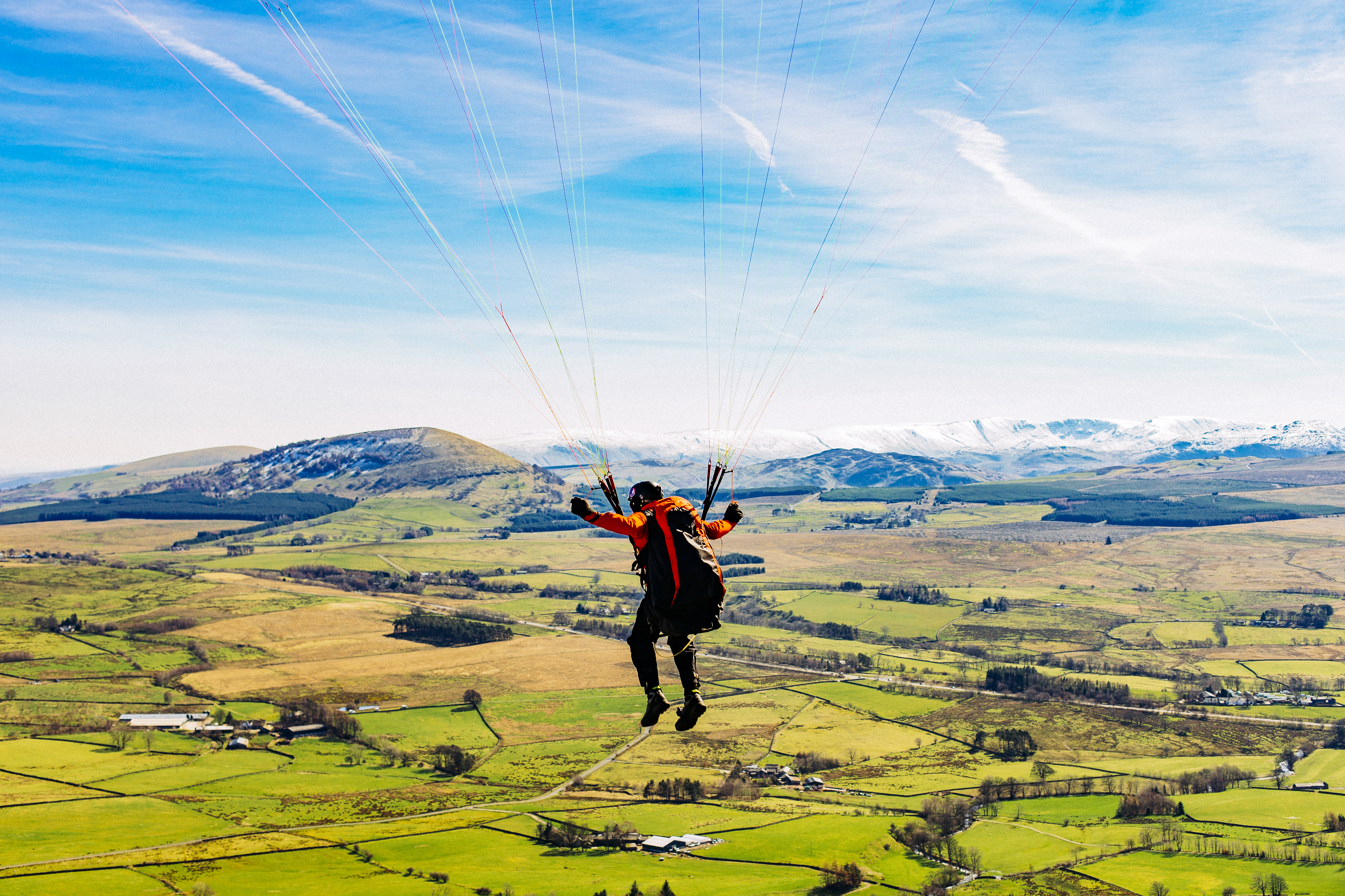 Person paragliding over a patchwork of green fields, with rolling hills and distant snow-capped mountains under a bright blue sky.