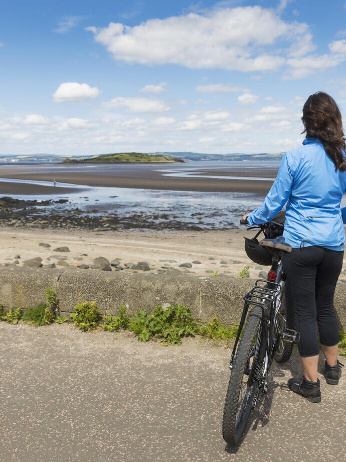 A female cyclist holding a bike and looking out over a beach.
