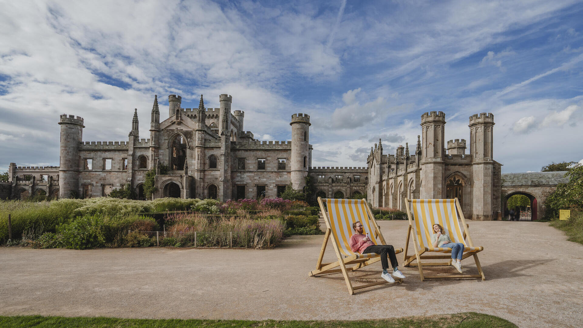 Zwei Personen sitzen auf großen Liegestühlen vor einem historischen britischen Schloss unter teilweise bewölktem Himmel.