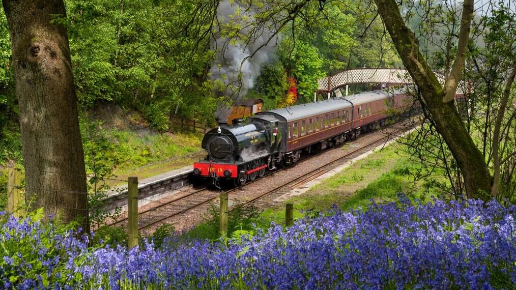 A steam train with lots of steam coming out, pulling at least three vintage carriages, gets ready to depart a small traditional railway station.