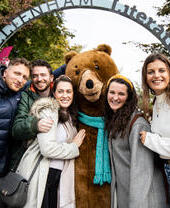 Menschen posieren mit einem großen Bärenmaskottchen unter einem Bogenschild mit der Aufschrift „Cheltenham Literature“