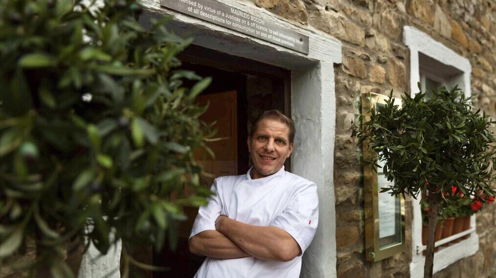 Chef in white uniform stands smiling at the entrance of a rustic stone building with white window frames and potted plants.