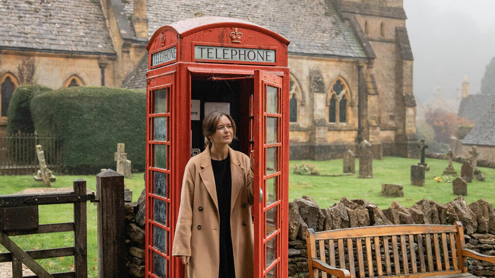 امرأة تغادر كشك هاتف أحمر تقليدي في قرية إنجليزية مع كنيسة حجرية في الخلفية.