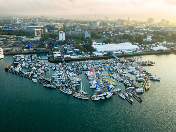 Aerial view of a marina filled with boats and yachts by the waterfront. Modern cityscape and event tents are visible in the background.