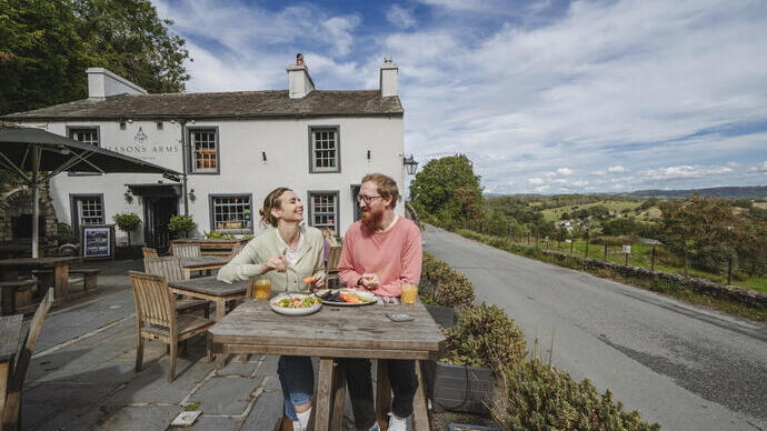 Two people dine outdoors at a rustic pub with countryside views, wooden tables, and a white building under a blue sky.
