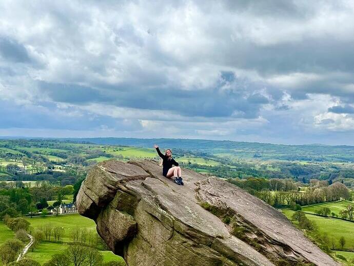 Person sitzt auf einem großen Felsen mit Blick auf eine grüne Landschaft unter bewölktem Himmel.
