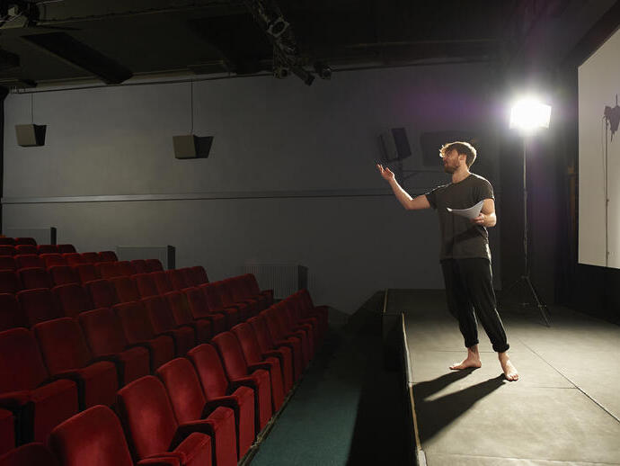 Un actor ensaya sus líneas en un escenario iluminado por un foco en un pequeño teatro comunitario.