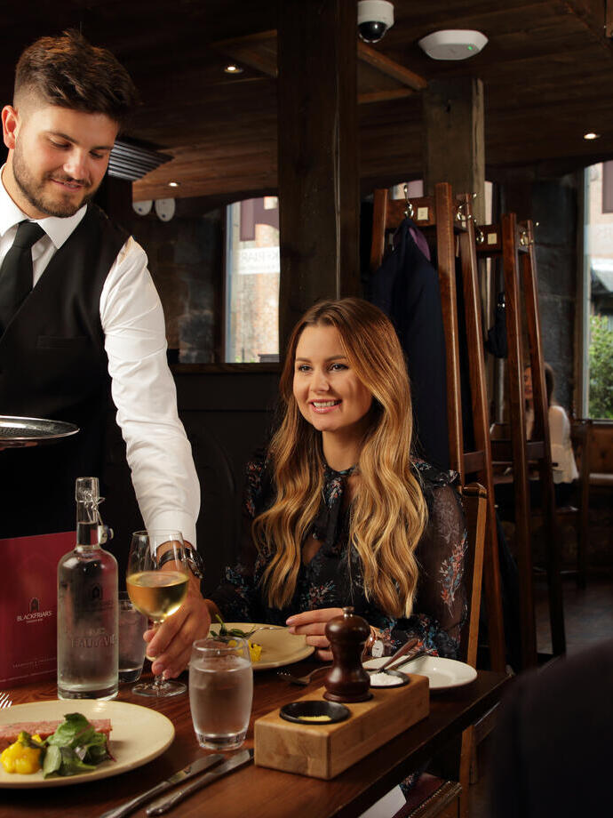 A smartly dressed staff member serving wine and food to a couple in a dining room 