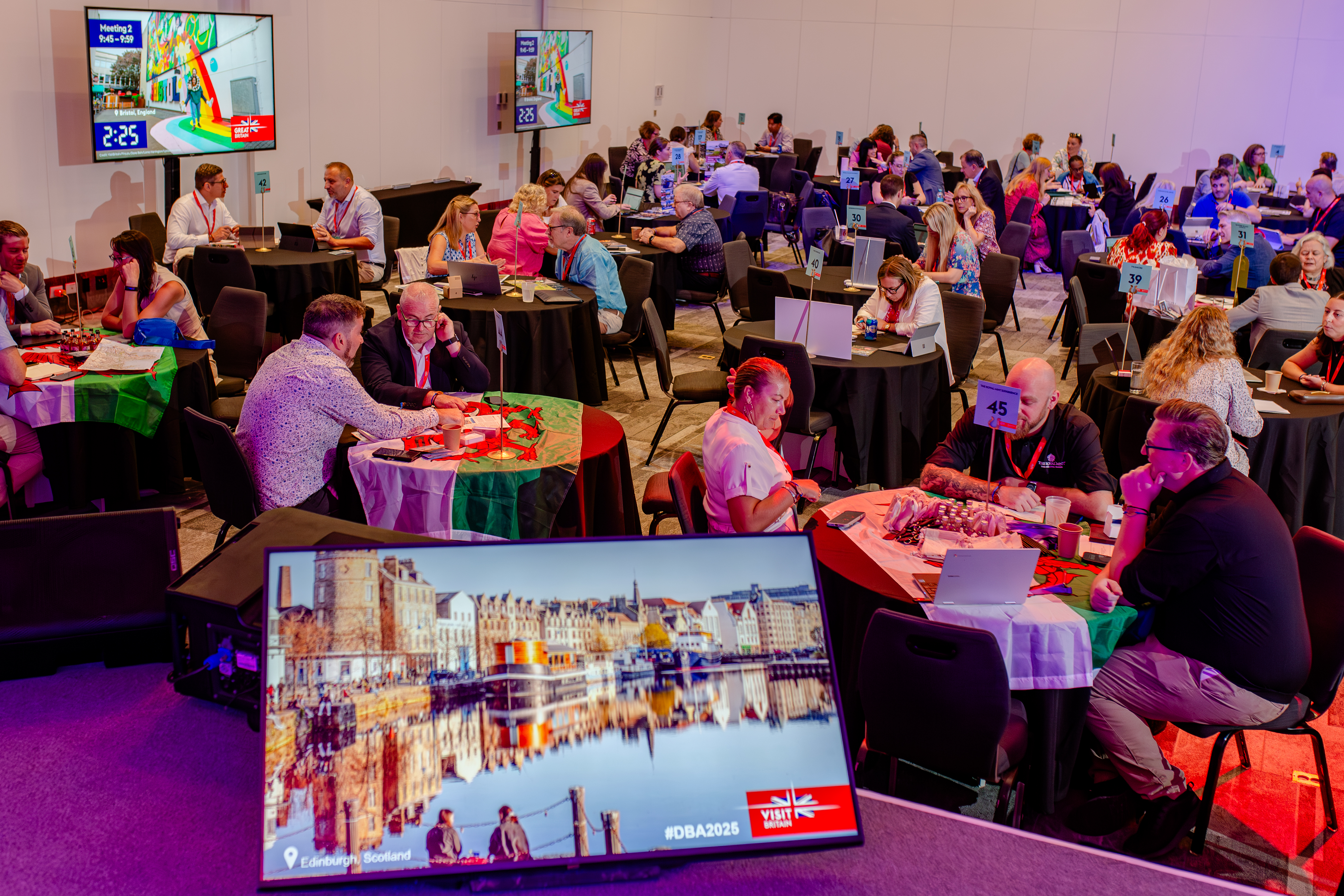 A conference room with round tables, people networking, laptops open, and a screen showing an image of Edinburgh, Scotland. Professional event atmosphere.