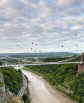 Hot air balloons floating above a river and a suspension bridge.