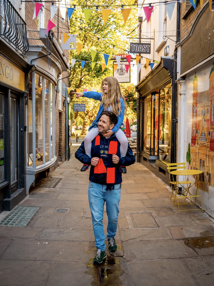 Ein Mann mit seinem Kind auf den Schultern geht durch eine Fußgängerzone mit Geschäften zu beiden Seiten
