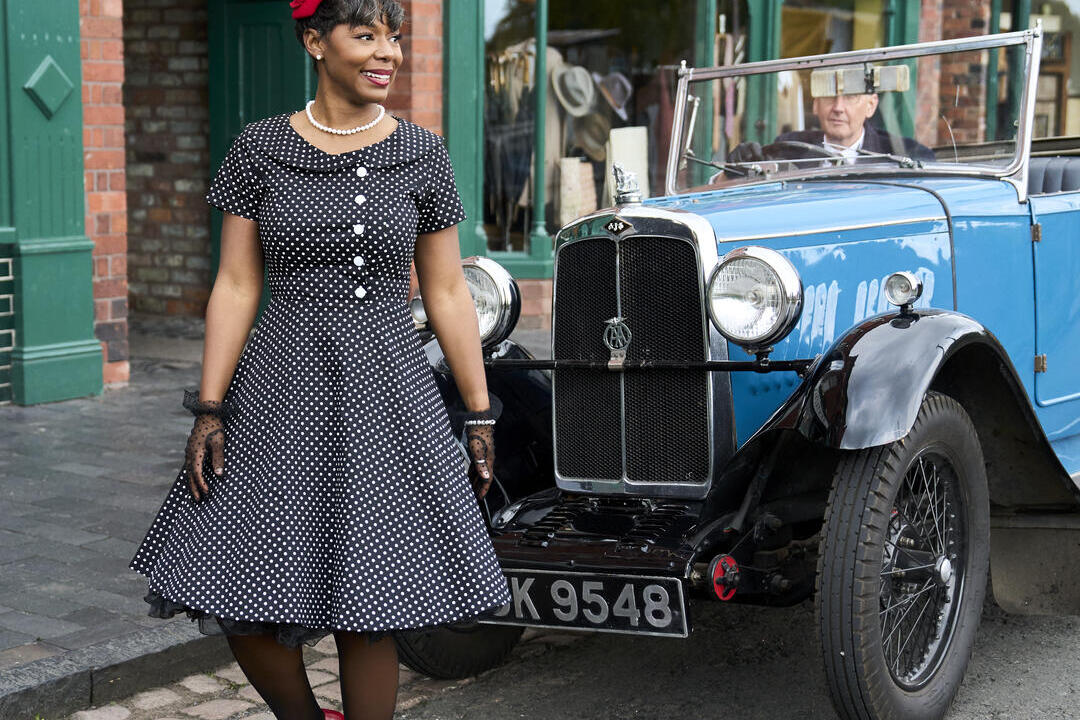 Une femme en tenue vintage se tient devant une voiture bleue classique, garée dans une rue bordée de bâtiments en brique d'époque.