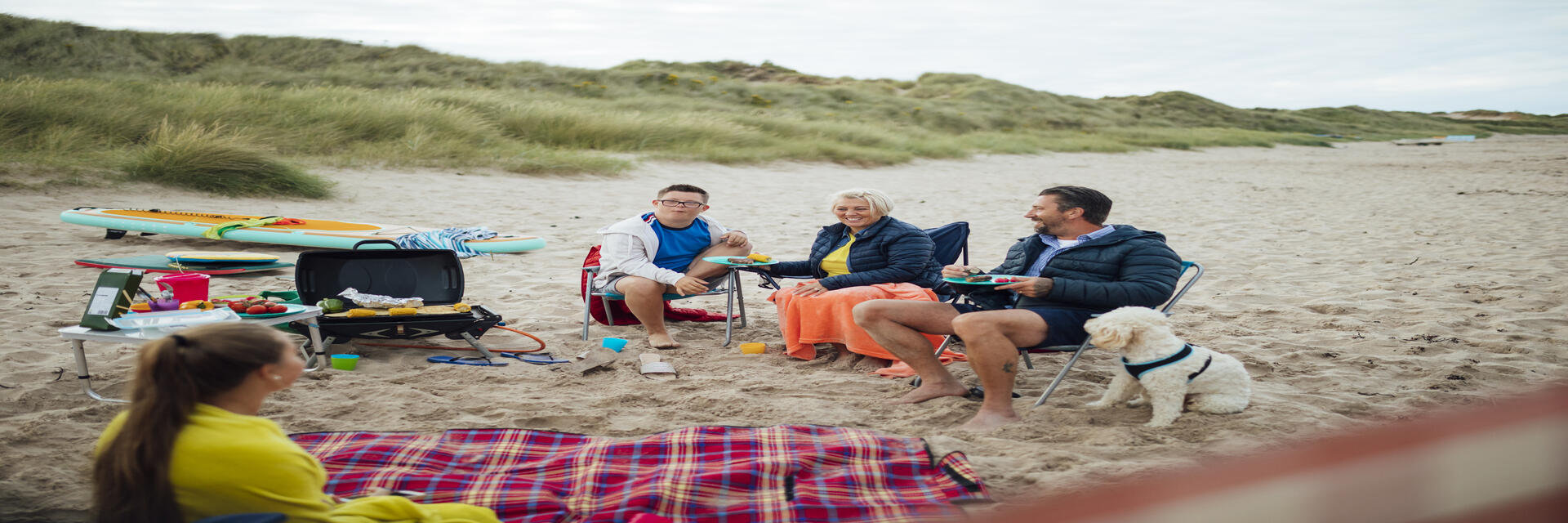 Eine Familie und ihr Hund sitzen neben einer Picknickdecke am Strand