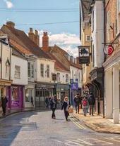 Rue commerçante juste après une averse. Des bâtiments historiques bordent la rue et les acheteurs marchent entre eux. Un ciel nuageux au-dessus.