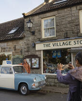 Street view of two people and a vintage police car outside a stone village store and coffee shop, with gift shop signs.