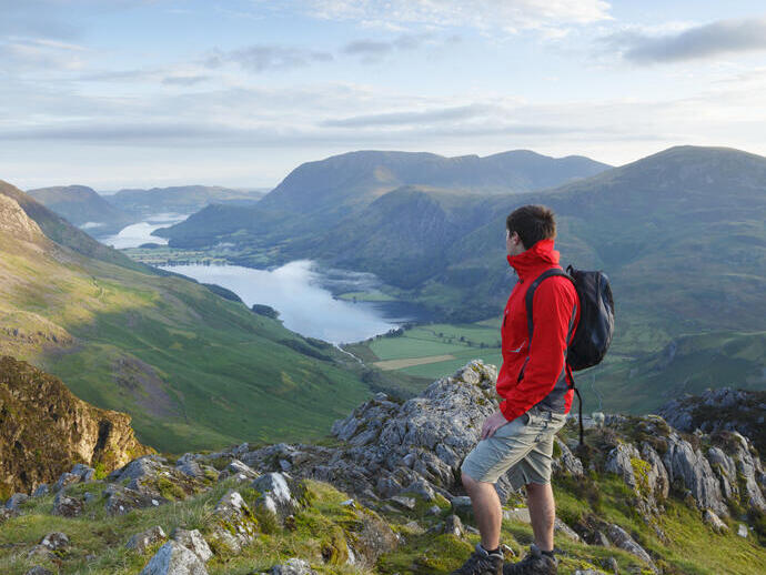 Ein Mann mit Rucksack steht auf einem Berg und blickt auf Hügel und einen See