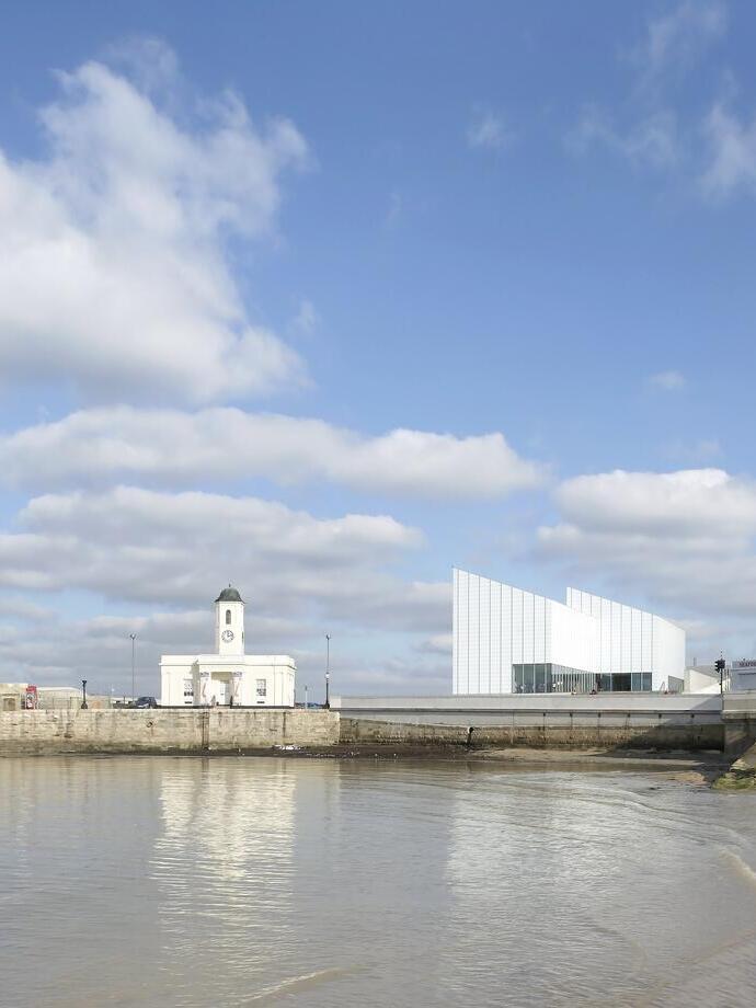 Modern building overlooking a harbour and beach, a row of townhouses in the background