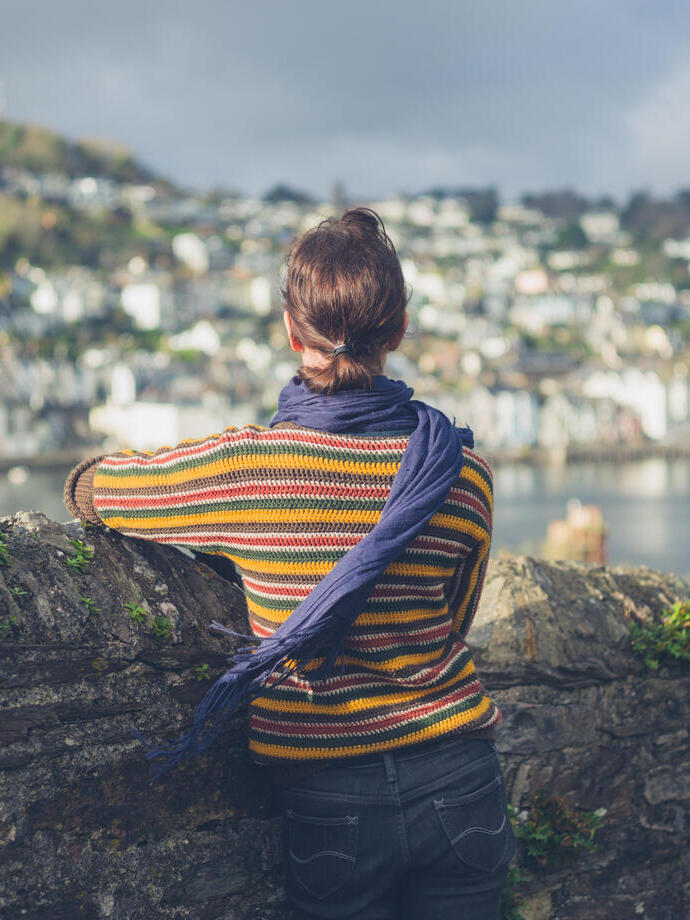 A young woman is standing by a stone wall in a village and is looking at the boats moored in the estuary