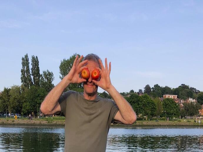 A person stands by the water holding two apples in front of their eyes, with trees and houses in the background.