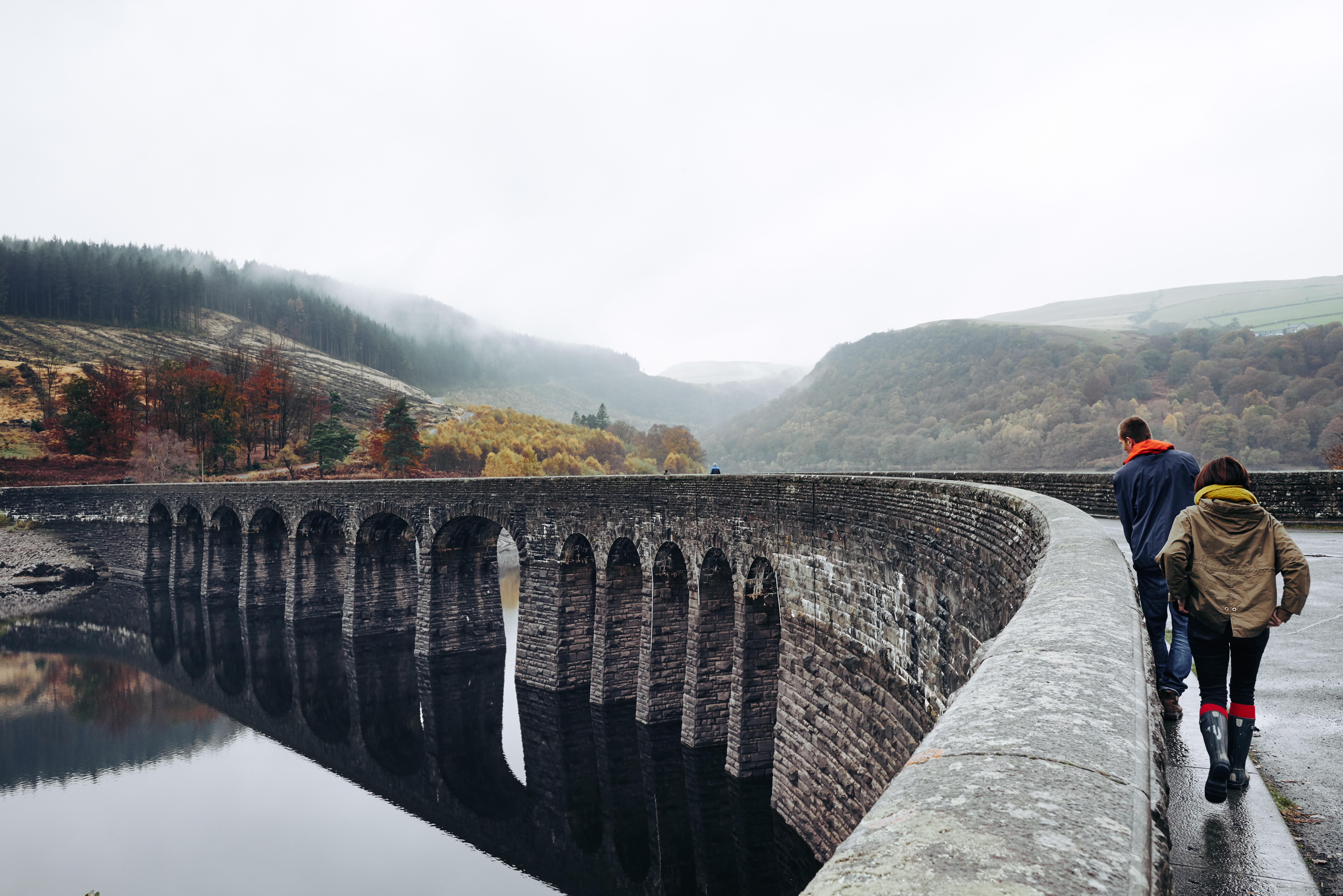 Two people walking on an arched bridge spanning a lake, with a valley in the background