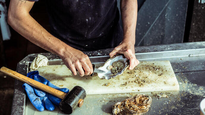 Person shucking an oyster on a cutting board with a mallet, gloves, and oyster shells on a metal counter.