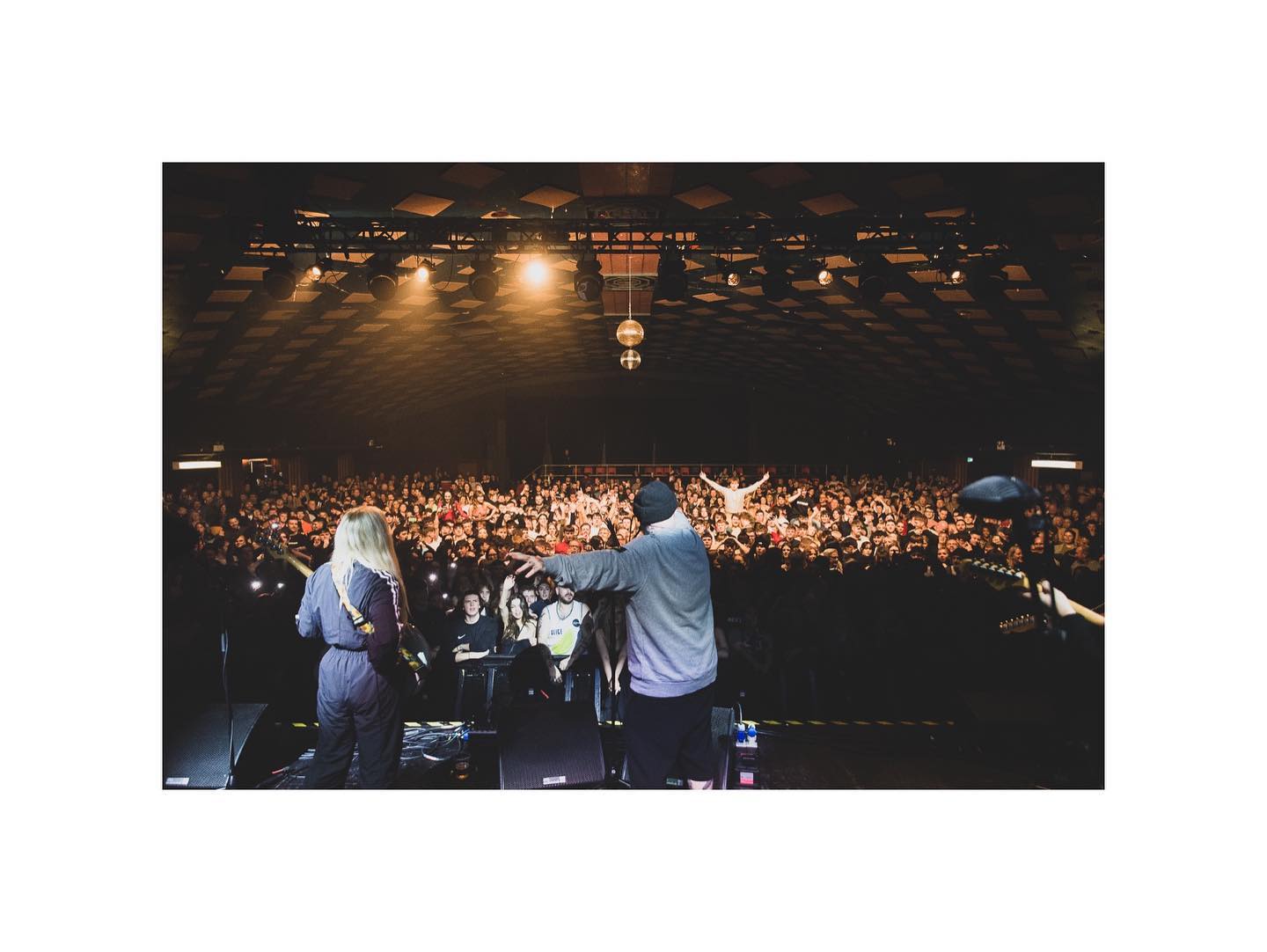 Two musicians on stage looking out to a large crowd of people dancing to the music and under bright lights and discoballs at Barrowland Ballroom in Glasgow