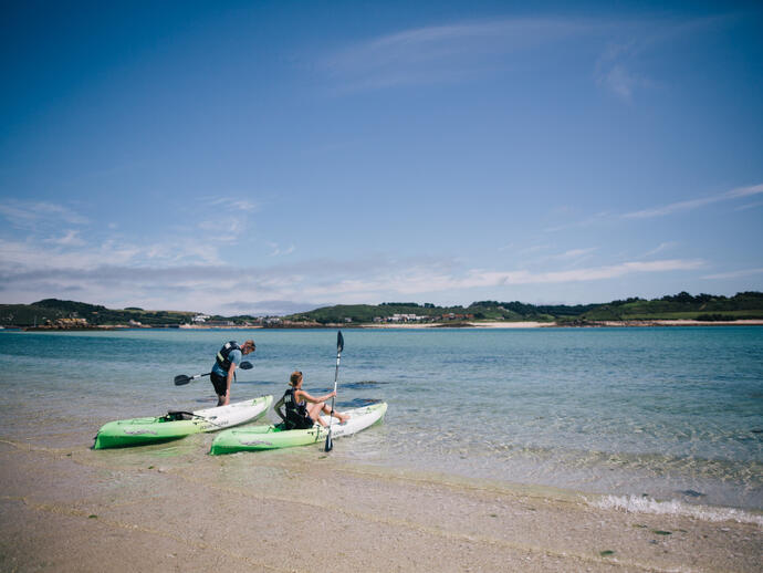 Two people with kayaks on a sandy shore beside clear blue water, with green hills in the background under a blue sky.