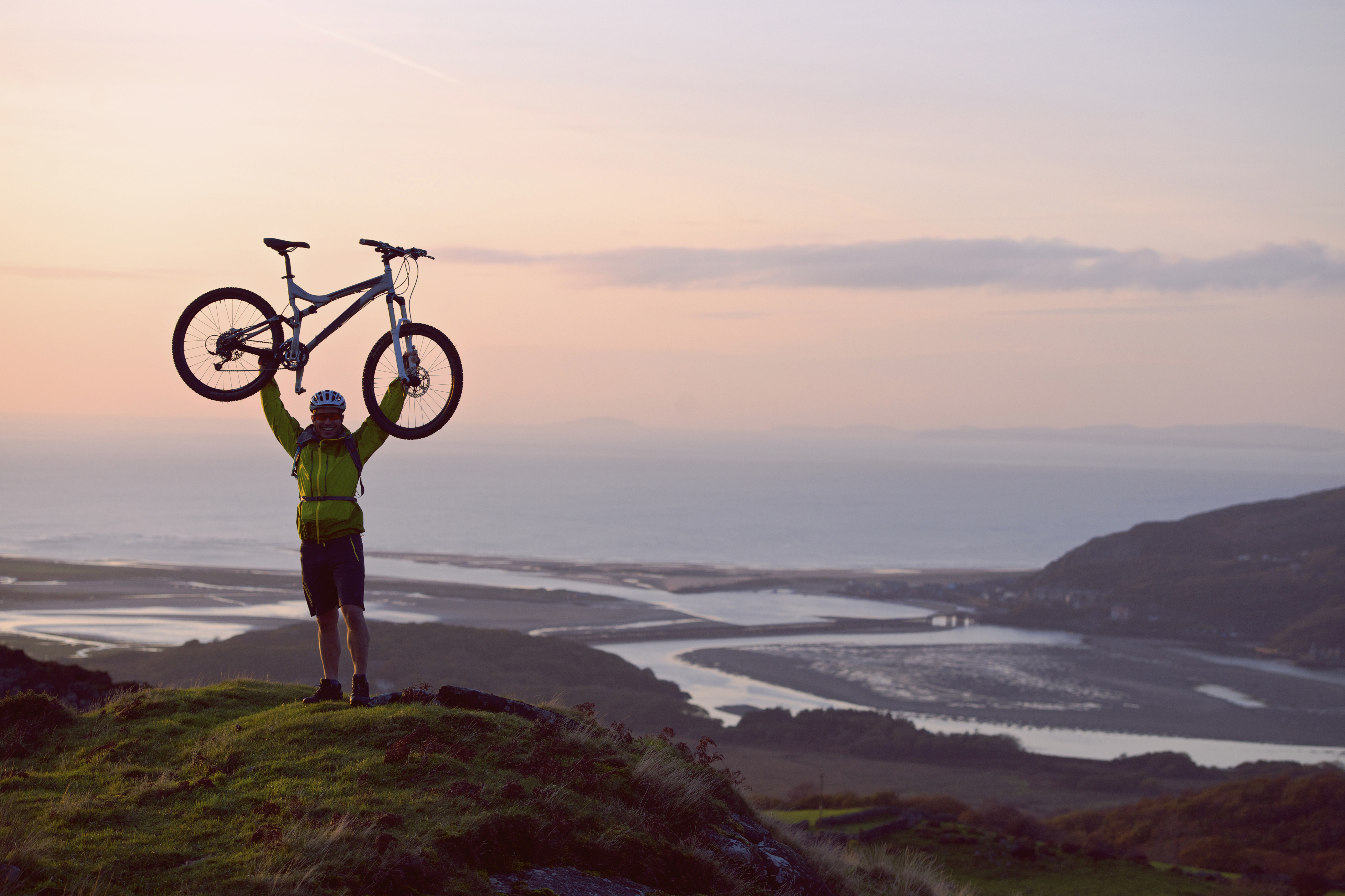 Un homme brandissant son VTT au-dessus de sa tête pour célébrer son arrivée au sommet d'une colline, avec la côte en contrebas