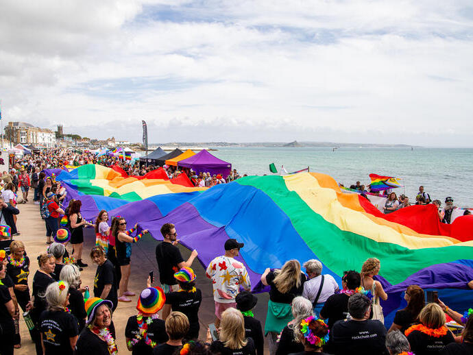 A crowd of people holding a large Pride flag on the coast in Cornwall