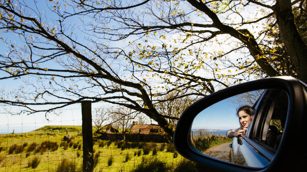 A countryside scene showing a view out of a car window. In the car mirror, a woman leans out enjoying the view of a rural road, green fields, trees and a clear blue sky.