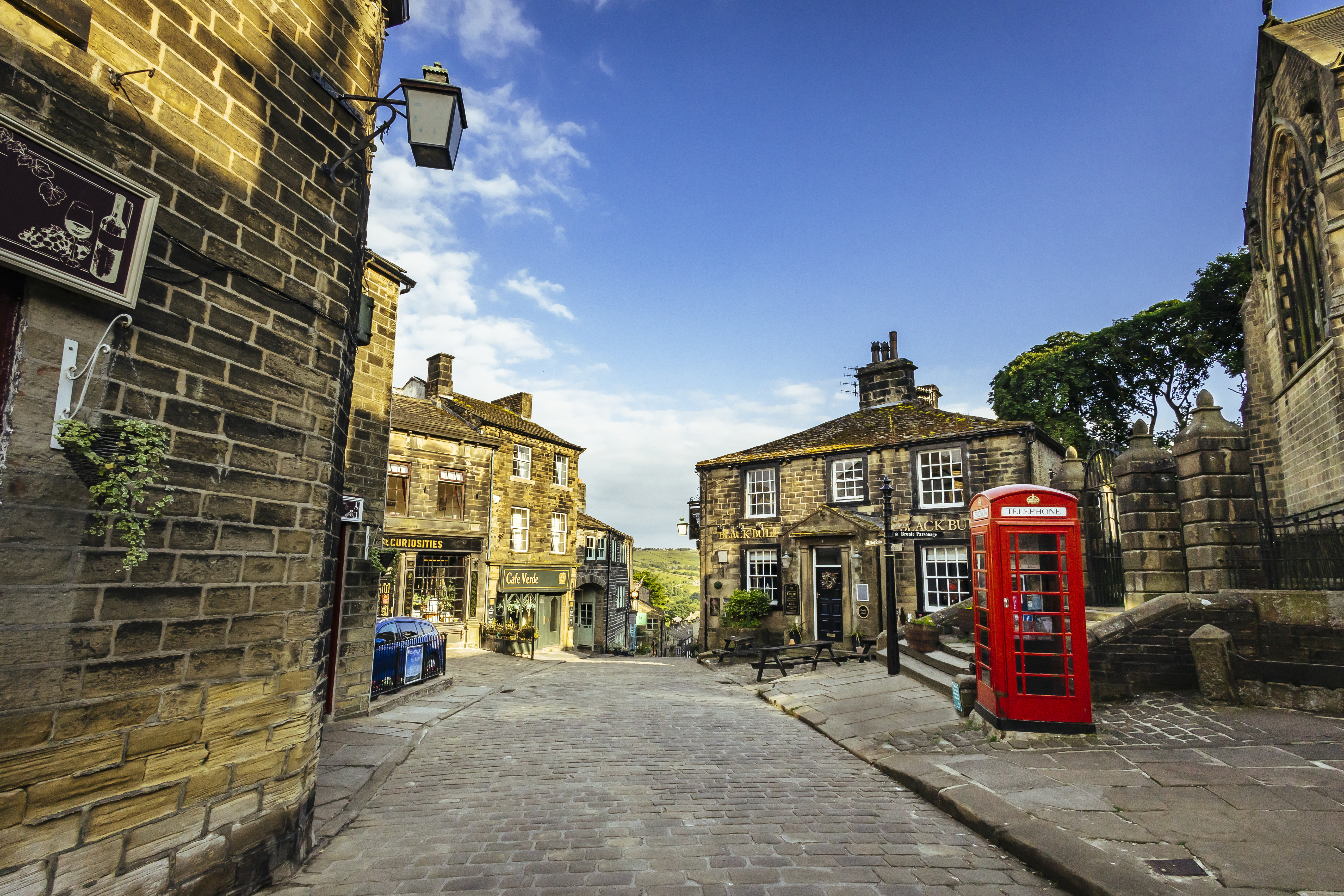 Stone street in a British village, historic stone buildings, red phone booth, blue sky, and a small café visible.