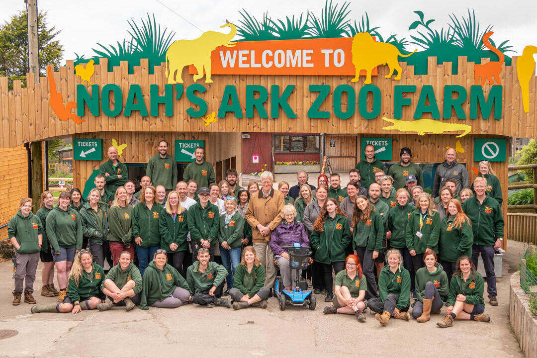 A sign which reads "Welcome to Noah's Ark Zoo Farm" with various staff members standing in front wearing matching branded tops. Noahs Ark Zoo Farm - Gold award winner for the Accessible and Inclusive Tourism Award at the VisitEngland Awards for Excellence 2023.