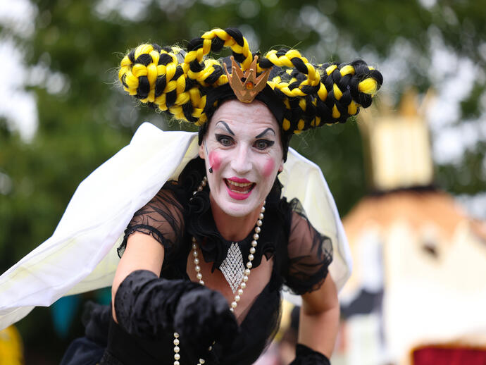 Person in elaborate costume and dramatic makeup with black and yellow braided hair outdoors, trees and event scenery in the background.