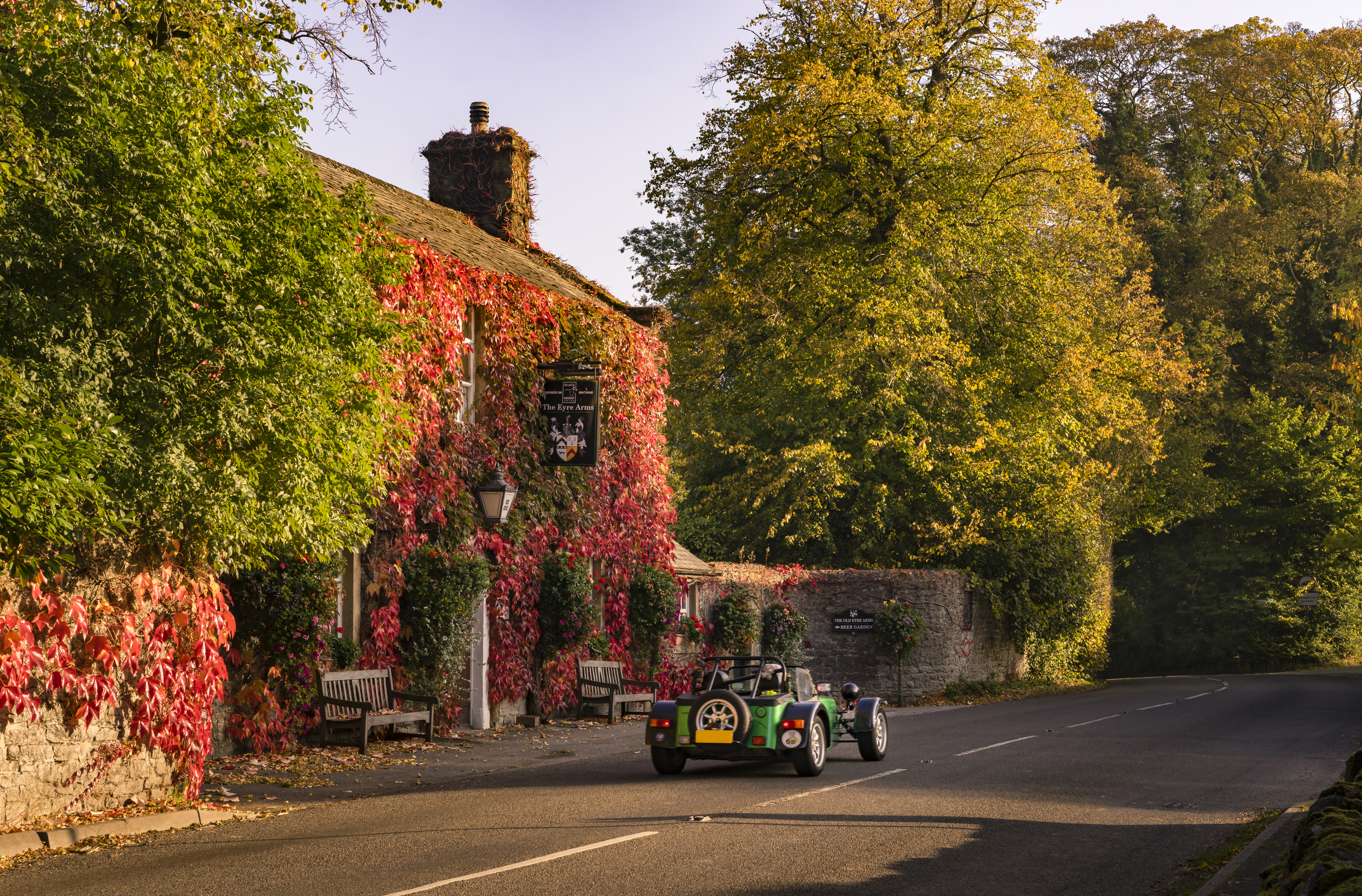 A classic car drives past a pub covered with ivy leaves