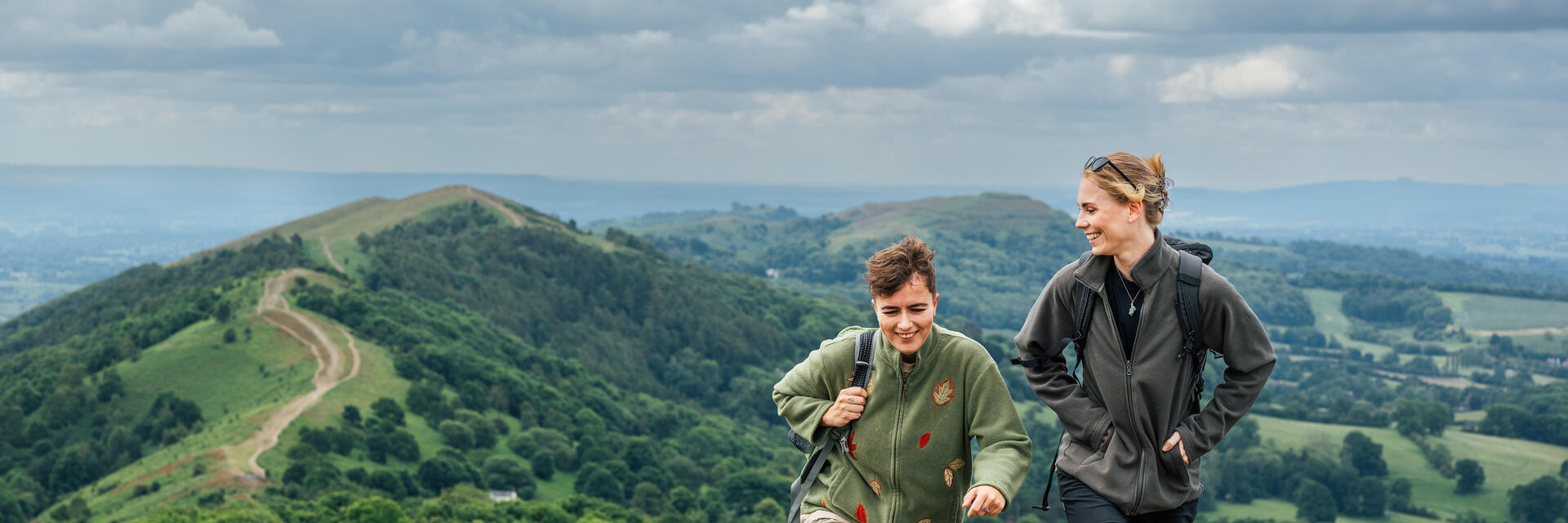 Two women hike carrying rucksacks along green hills on a cloudy day