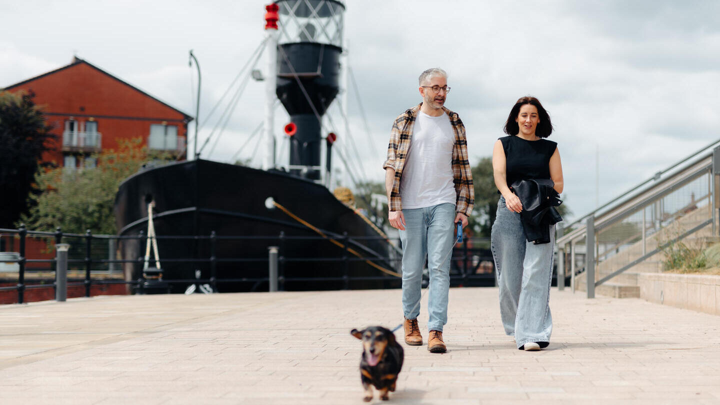 Two people walking a small dog along a paved waterfront with a historic ship and residential buildings in the background.