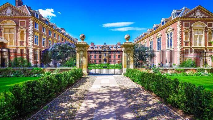 Pathway leading to ornate gates and historical brick buildings, with manicured gardens and bright blue sky in the background.