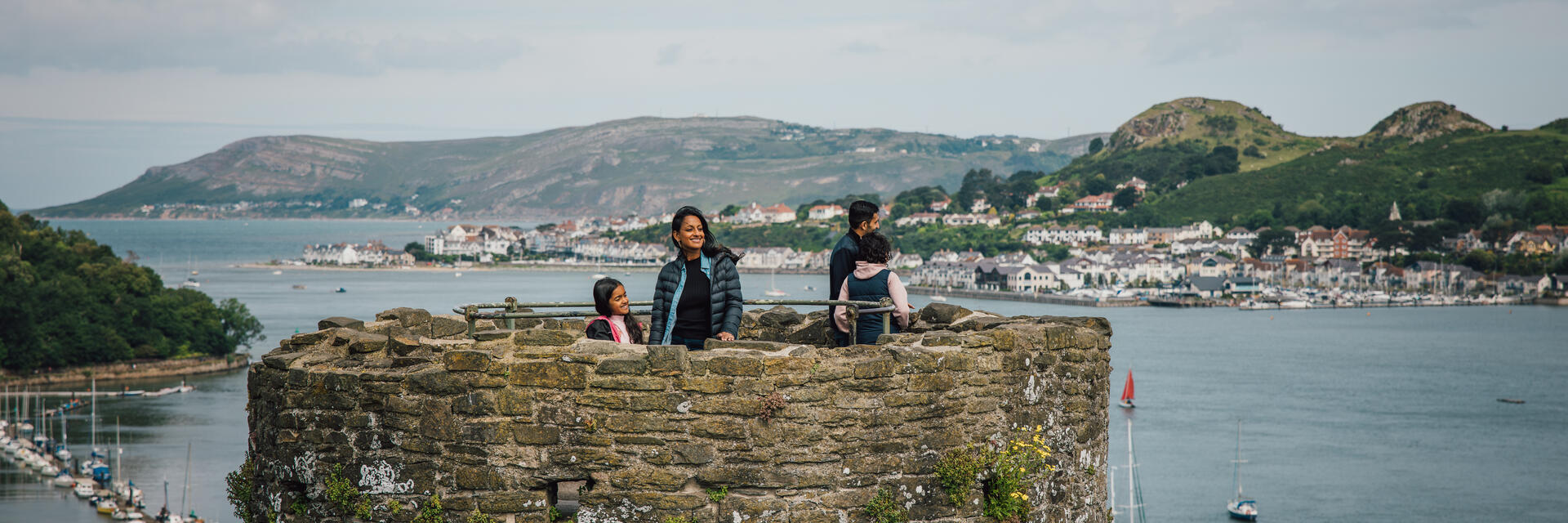 People standing atop an old stone castle tower overlooking water, sailboats, and a coastal town with hills in the background under a cloudy sky.