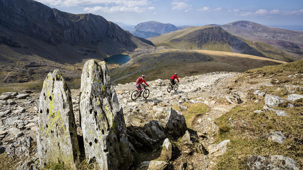 Dos ciclistas de montaña pedaleando por un camino pedregoso. Lago al frente