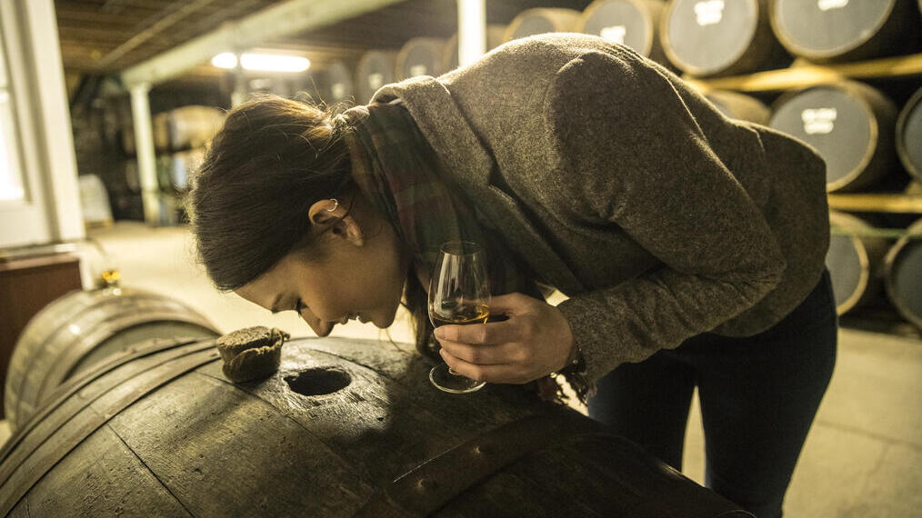 Woman in a distillery, holding a glass of whisky and looking through a hole in an oak barrel used for ageing whisky