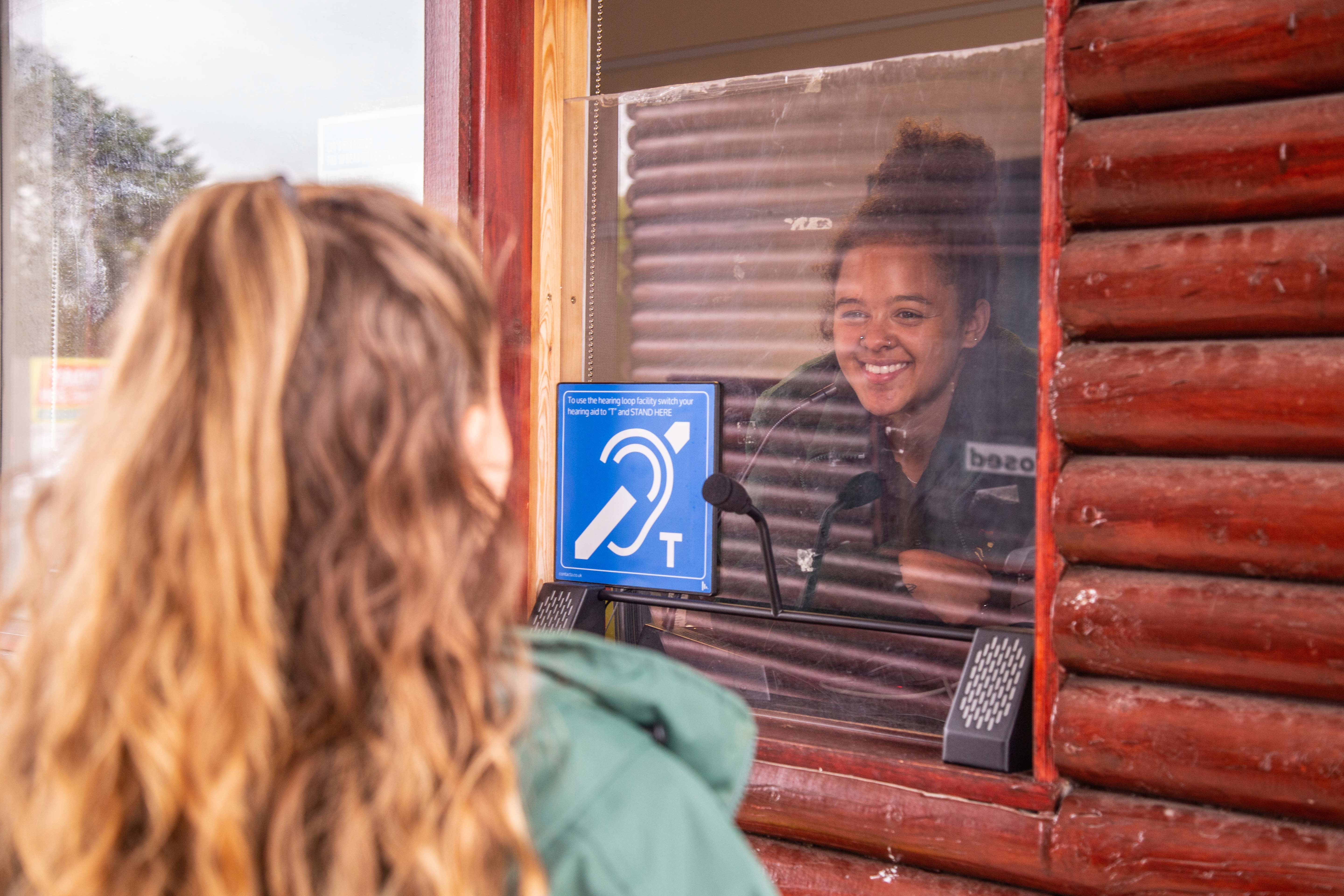 A person inside a ticket booth smiles at someone standing outside. A blue hearing loop sign is prominently displayed. Noahs Ark Zoo Farm - Gold award winner for the Accessible and Inclusive Tourism Award at the VisitEngland Awards for Excellence 2023.